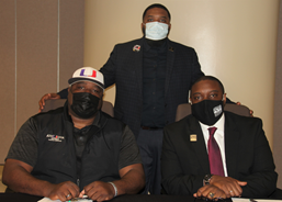 Three African American males wearing face masks and business attire posing for a photo; two males are sitting, 一只雄性站着, both males who are sitting have their hands clasped in front of them on a table.